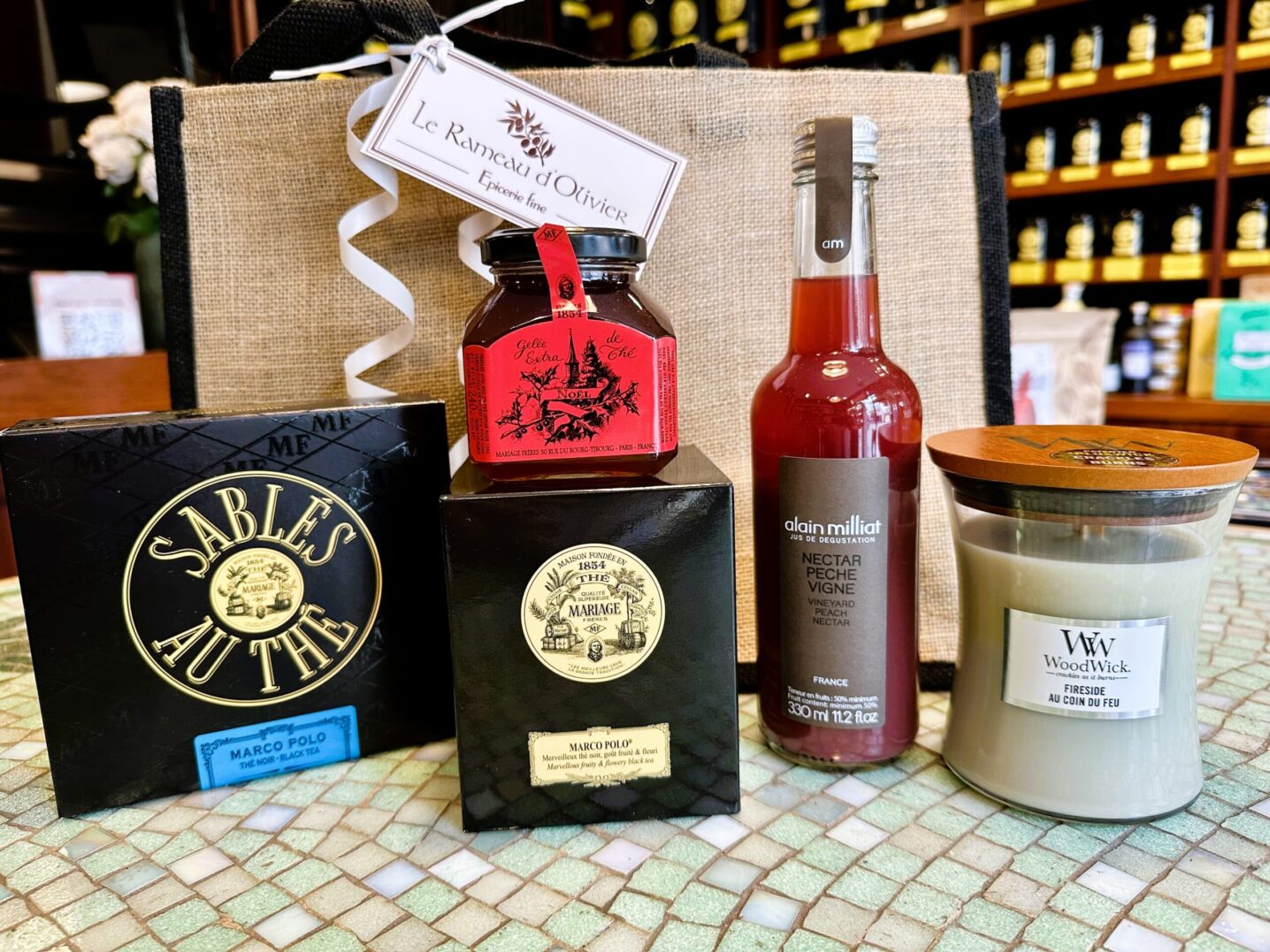 Une Sélection Pause Goûter un thé Marco Polo, des sablés au thé et une gelée de Thé Mariage Frères, un jus de fruits Alain Milliat et une bougie Woodwick modèle petit sablier dans un élégant sac en jute
